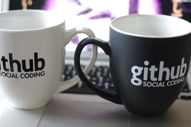 Две кружки GitHub рядом с клавиатурой на рабочем столе разработчика